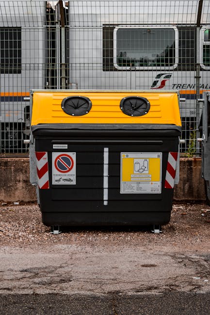A large outdoor waste container featuring a black body with a yellow lid, situated on a gravel surface adjacent to a low brick boundary wall. The container has two circular ventilation openings on the