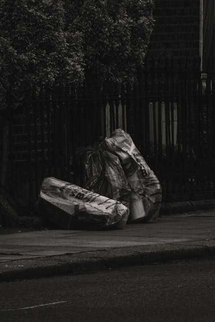 Three large black refuse bags made of plastic, filled with waste materials, are placed on the edge of a sidewalk beside a black metal fence and a paved road. The bags appear to be tightly knotted at t